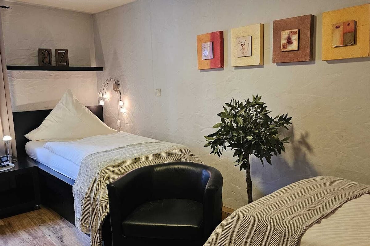 A bedroom with two beds, a nightstand with a lamp, a potted plant, and artwork on the wall.