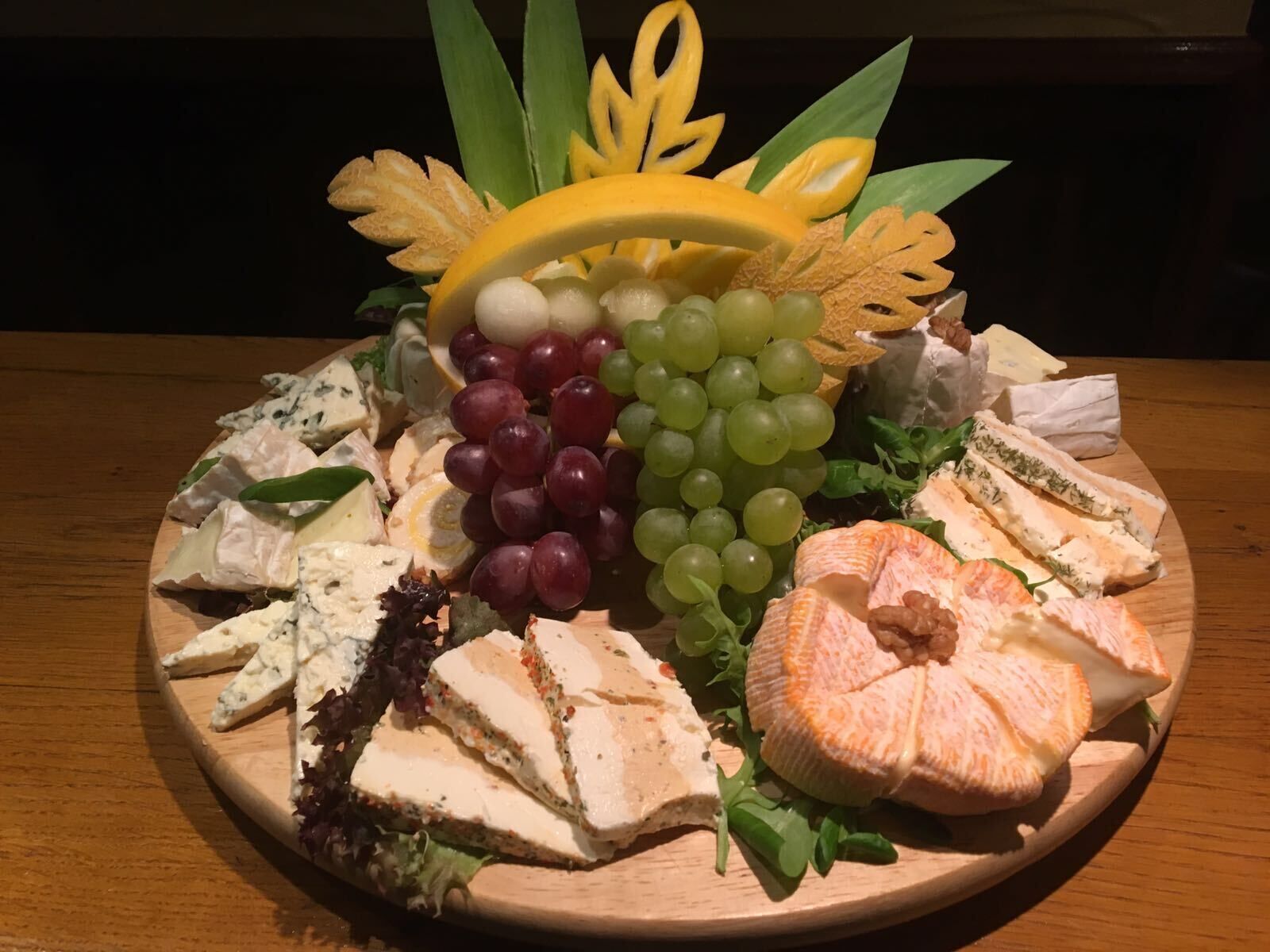 A wooden platter with various cheeses, grapes, and leafy greens.