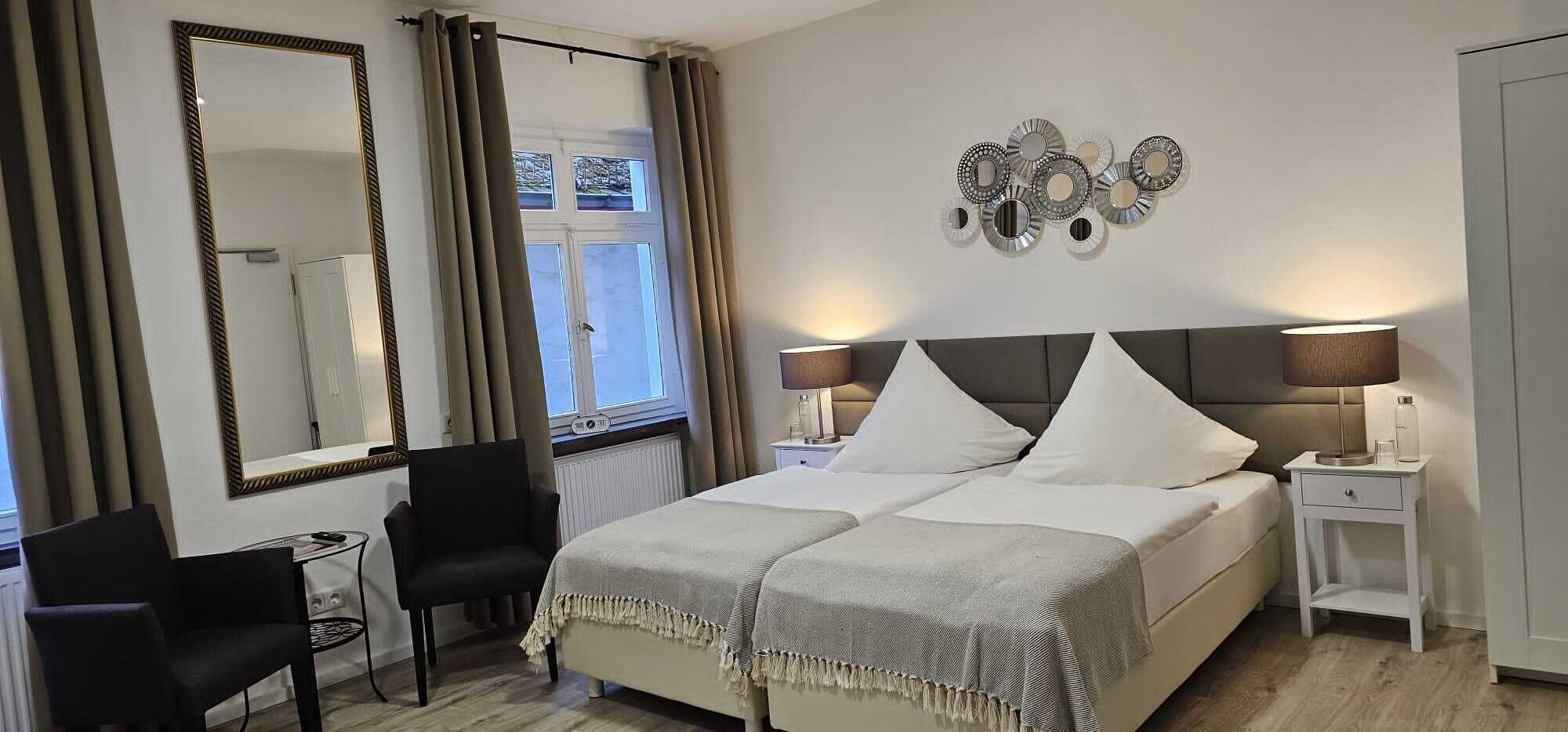 Una habitación de hotel con una cama grande, dos mesitas de noche con lámparas, una silla, un escritorio, un espejo y una ventana con cortinas.