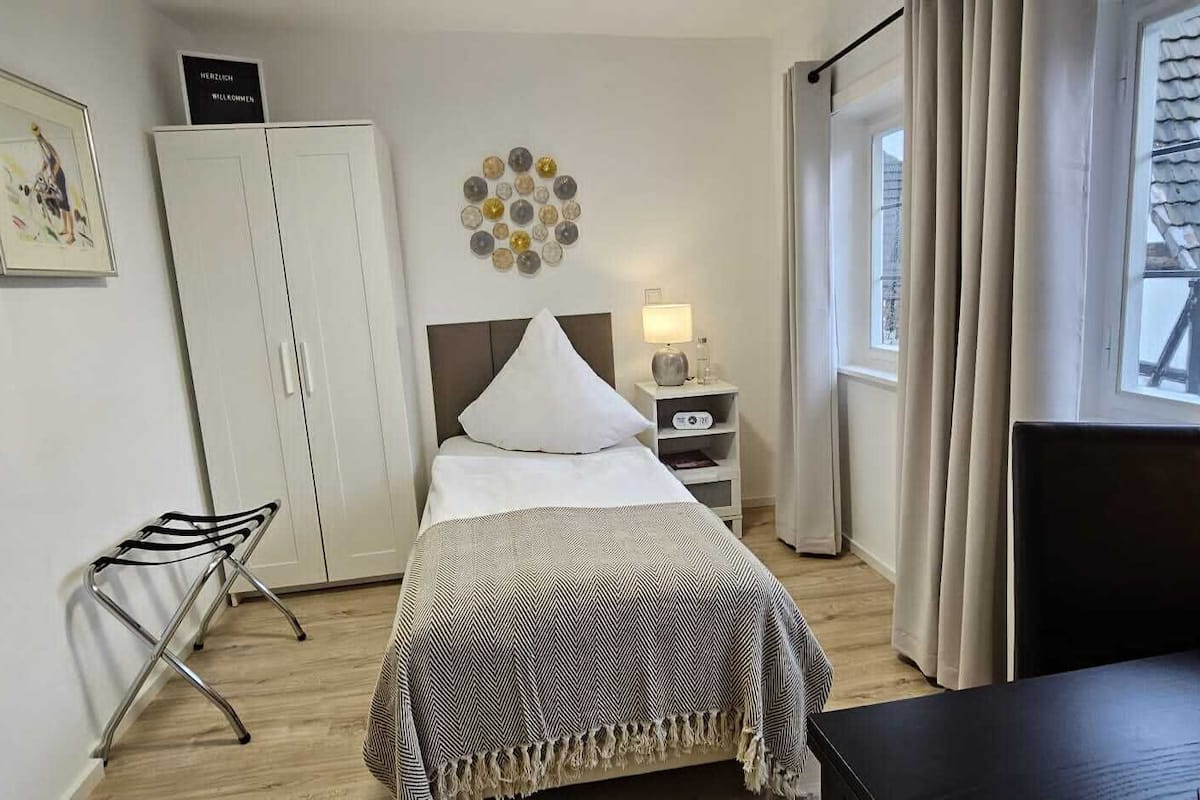 A hotel room with a bed, a bedside table with a lamp, a wardrobe, a chair, and a window with curtains.