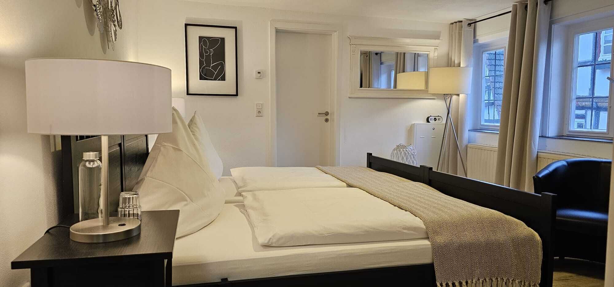 A hotel room with a bed, a bedside table with a lamp, a mirror, and a window with curtains.