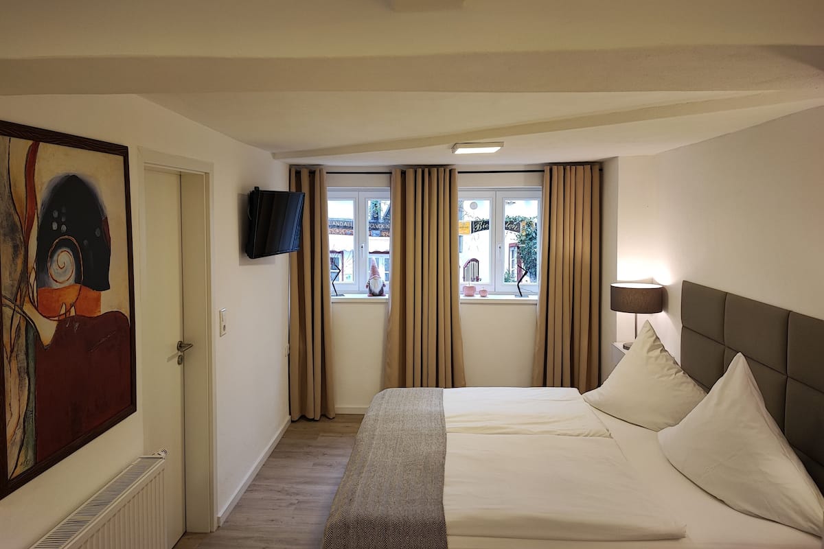 A hotel room with a bed, a TV mounted on the wall, a window with curtains, and a painting on the wall.