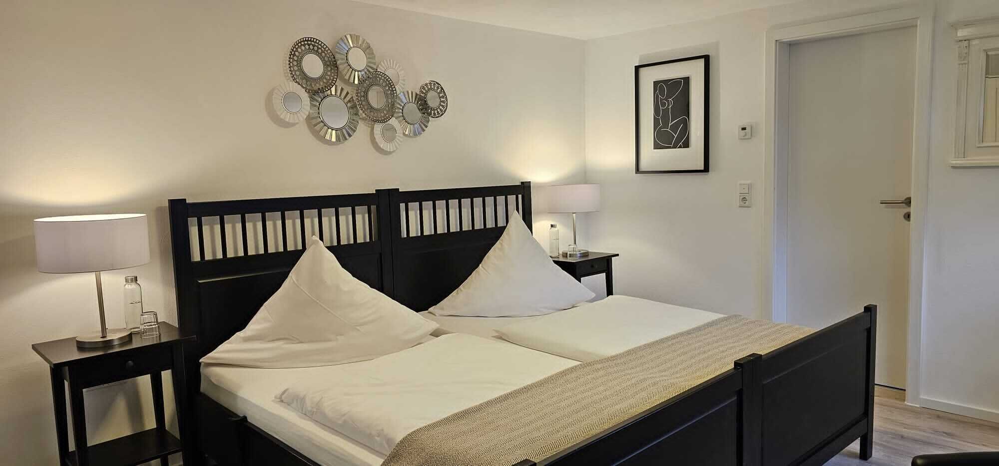 A neatly made bed with white linens and pillows, a bedside table with a lamp, and a wall with decorative mirrors and a framed picture.