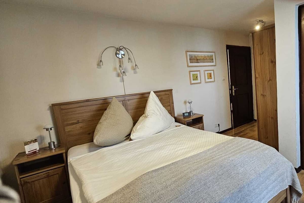 A bedroom with a bed, wooden headboard, bedside tables, a wall-mounted lamp, and a wooden wardrobe.