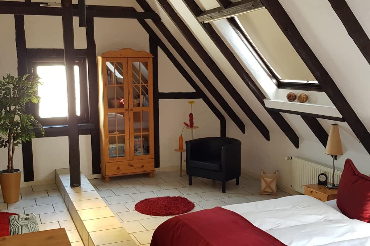 A bedroom with a sloped ceiling, a wooden cabinet, a black chair, a bed with red bedding, a red rug, and a potted plant.
