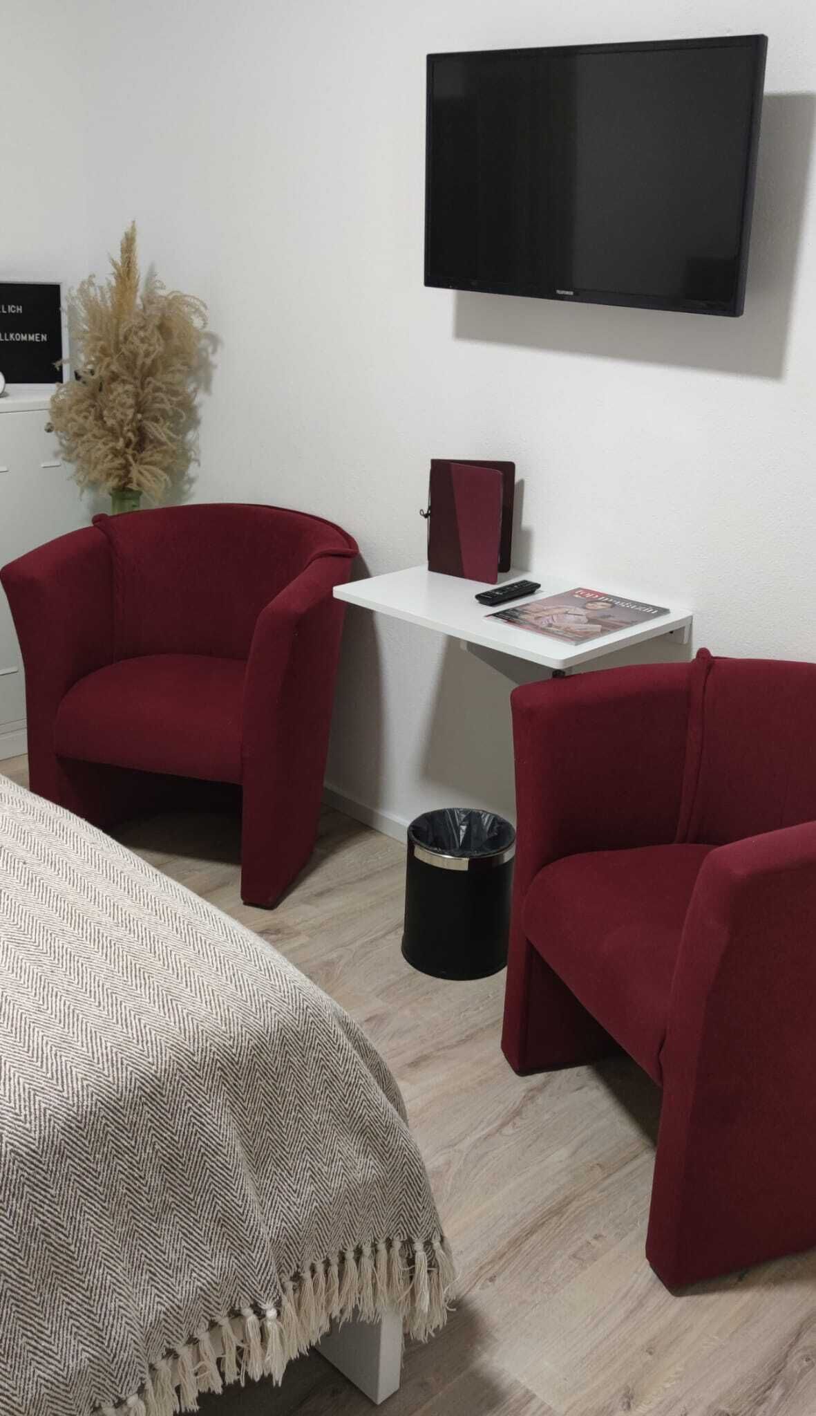 A room with a bed, two red armchairs, a small white table, a TV mounted on the wall, and a trash bin.