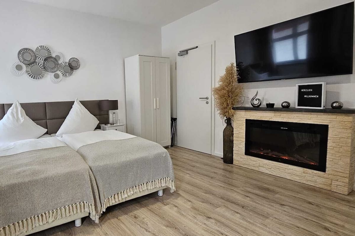 A modern bedroom with a bed, bedside tables, a TV, a fireplace, and a door.