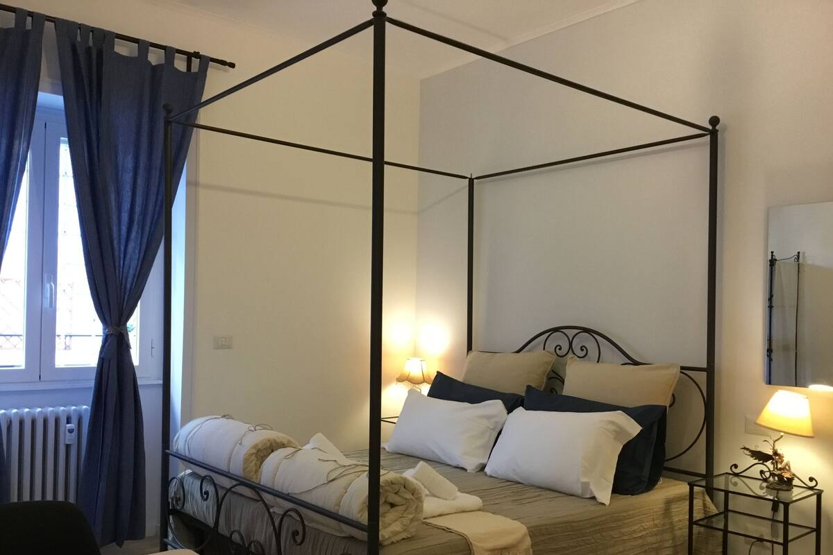 A four-poster bed with a headboard, a bedside table with a lamp, and a window with curtains.