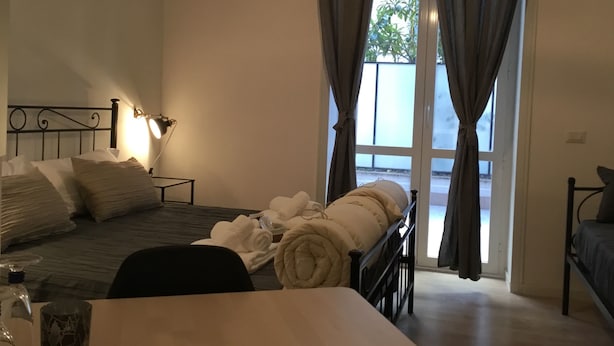 A bedroom with a bed, bedside table, lamp, and a view through a window with curtains. - Le Fate Suites (Rome)