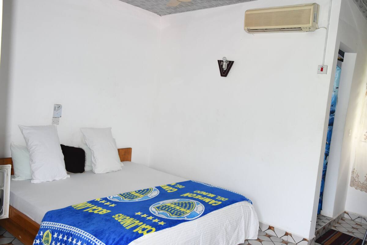 A bedroom with a bed, white walls, a ceiling fan, and a window with a curtain.