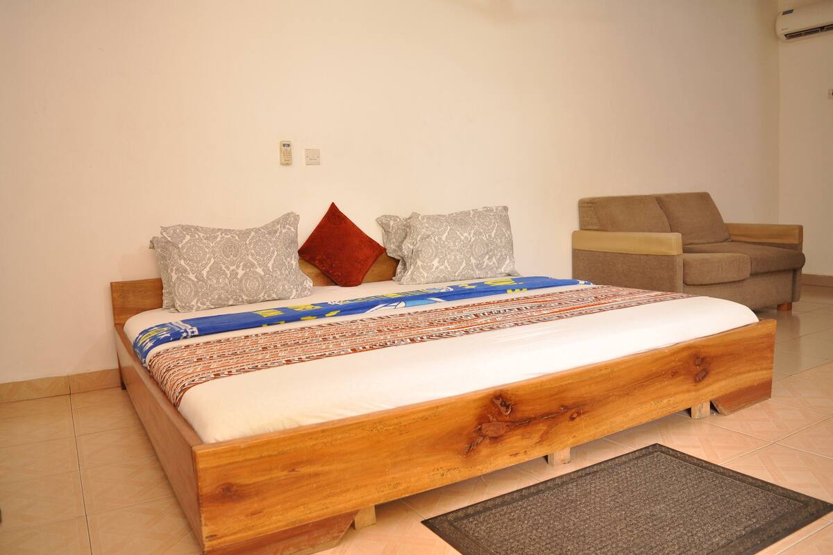 A bedroom with a wooden bed, patterned bedding, a red pillow, and a beige armchair.
