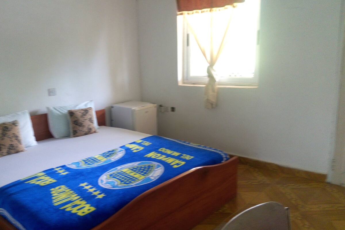 A small, simple room with a bed, a window with curtains, a radiator, and a small cabinet.