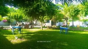 Spazio all'aperto con prato verdeggiante, due tavoli blu e sedie, circondato da alberi e un edificio bianco sullo sfondo.