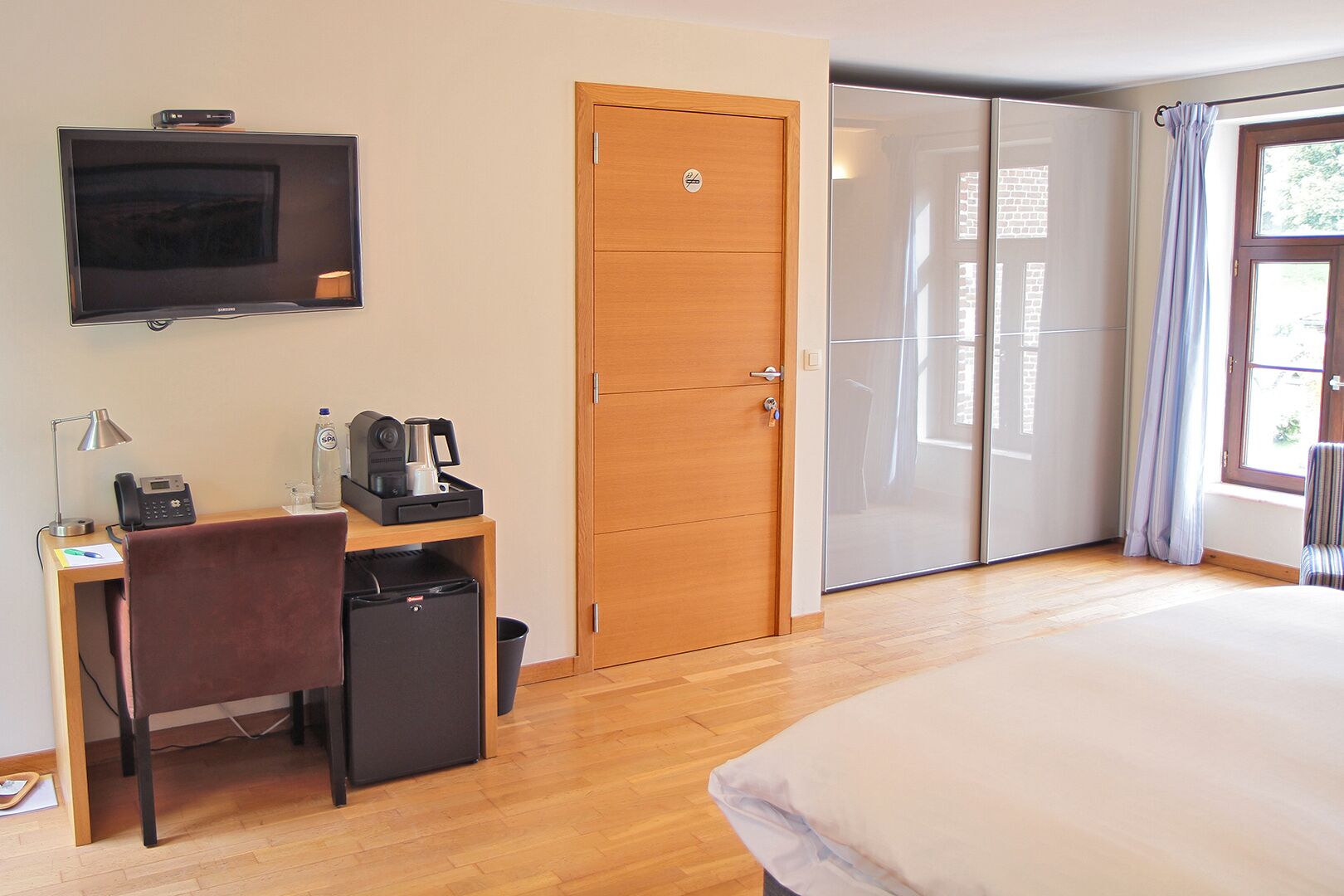 A hotel room with a bed, a desk with a chair, a TV, a coffee machine, and a wardrobe.