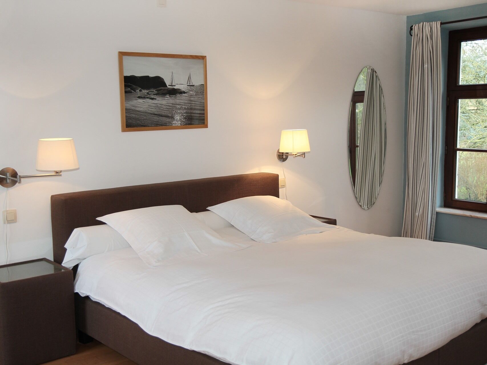 Una cama bien hecha con sábanas blancas, un cuadro enmarco de un paisaje costero, dos lámparas fijadas a la pared y una ventana con cortinas.