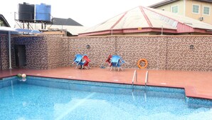A swimming pool with blue water, surrounded by a tiled deck. There are two blue lounge chairs and a red lounge chair. A lifebuoy is visible on the deck. - TommsVille Hotels (Uyo)