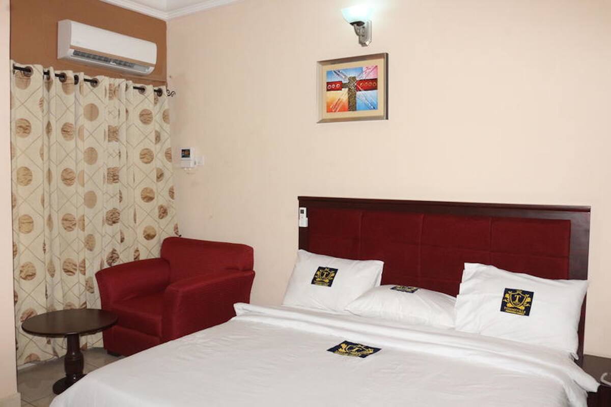 A hotel room with a bed, a red armchair, a small table, and a wall-mounted air conditioner.