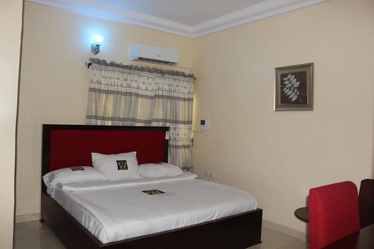 A hotel room with a bed, a red headboard, a wall-mounted air conditioner, a framed picture, and a small table with chairs.