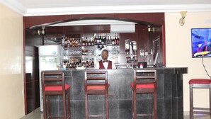 A bar area with a bartender, three wooden bar stools with red cushions, a well-stocked bar counter, and a television mounted on the wall. - TommsVille Hotels (Uyo)