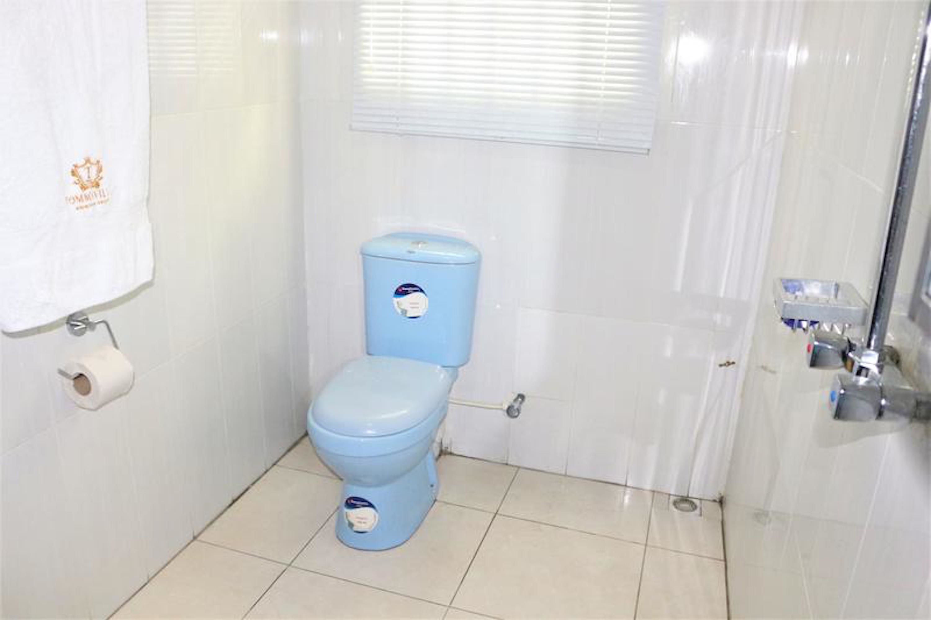 Um banheiro com vaso sanitário azul, uma toalha branca pendurada na parede e uma janela com persianas.