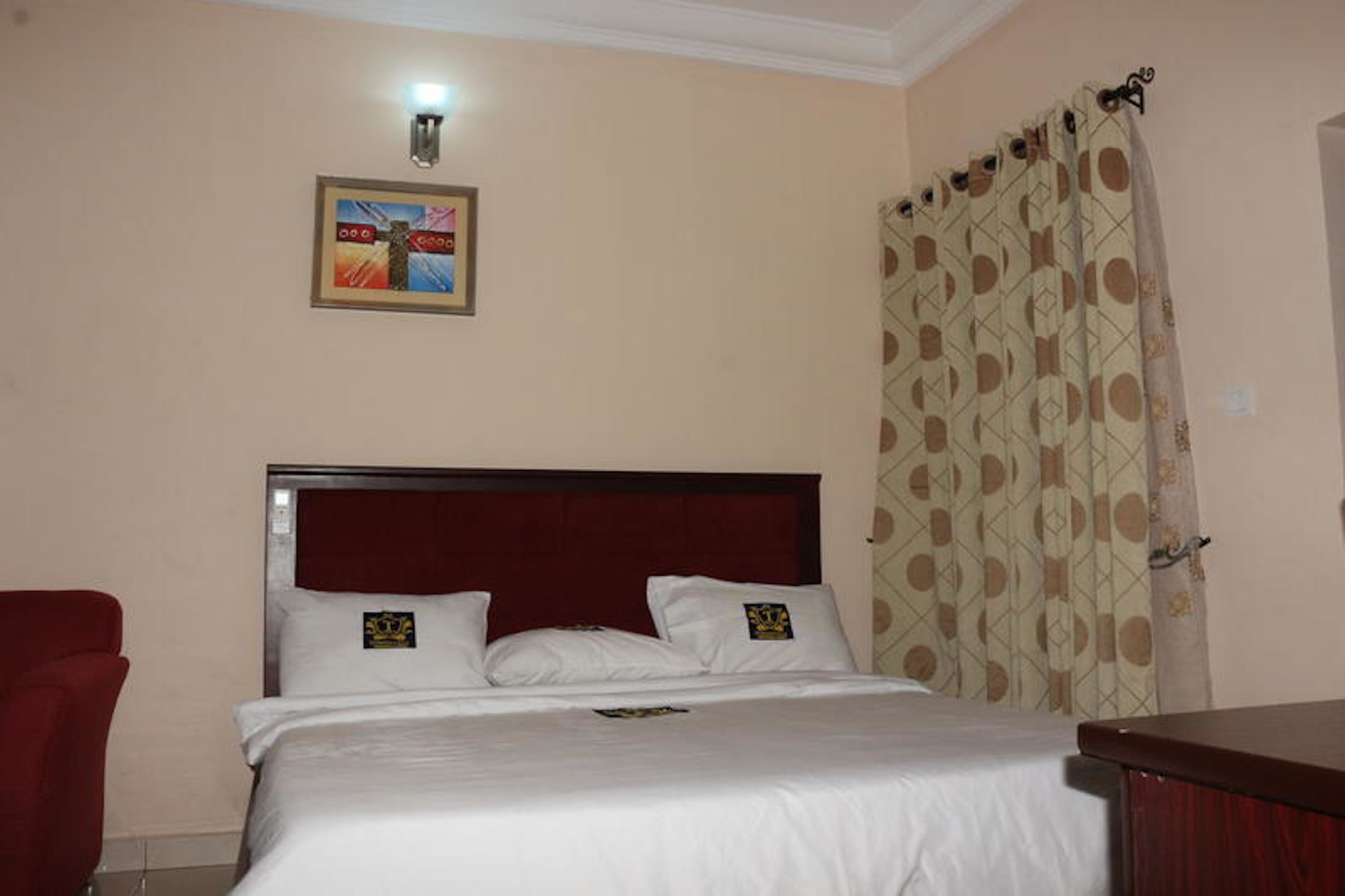 A hotel room with a bed, a red chair, a painting on the wall, and patterned curtains.