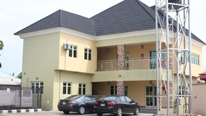 A two-story building with a balcony, a water tank on the side, and two cars parked in front. - TommsVille Hotels (Uyo)