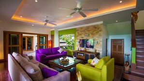 Un salon moderne avec un canapé, un fauteuil et une table basse. Un téléviseur est fixé au mur et un ventilateur de plafond est installé.