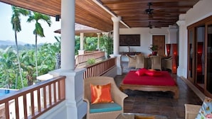 Un balcon spacieux doté d’un plafond en bois, d’un canapé rouge, de fauteuils en osier et d’une table en verre sur laquelle est posée une bougie.