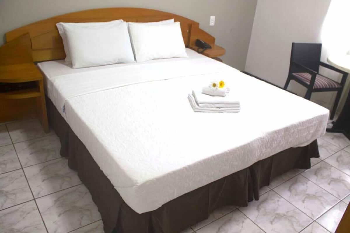 A neatly made bed with white linens, a small table with a chair, and a bedside table with a lamp.