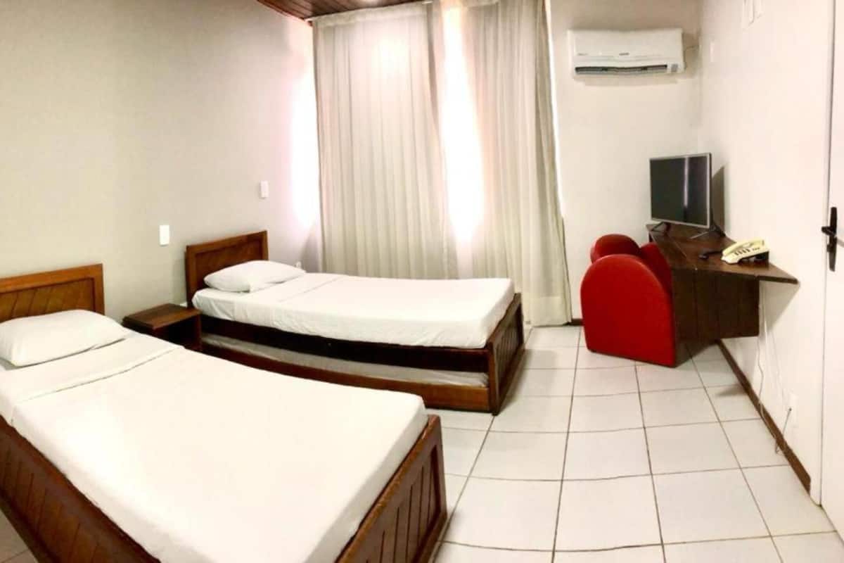 A room with two single beds, a desk with a chair, a television, and an air conditioning unit.