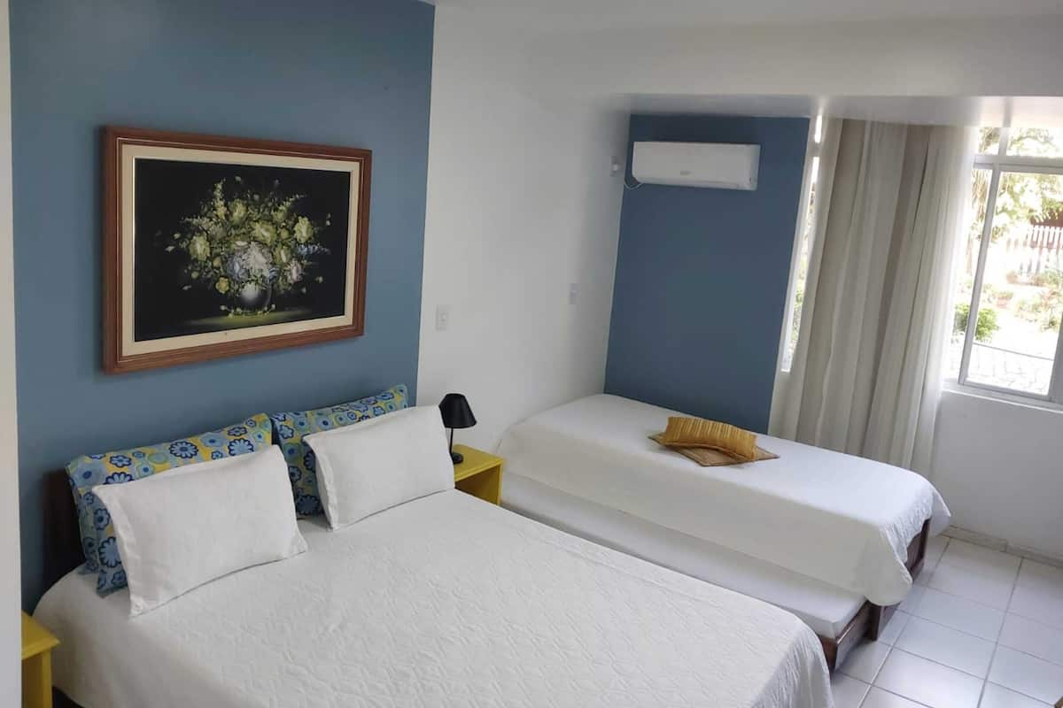 A hotel room with two beds, a painting on the wall, an air conditioner, and a window with curtains.