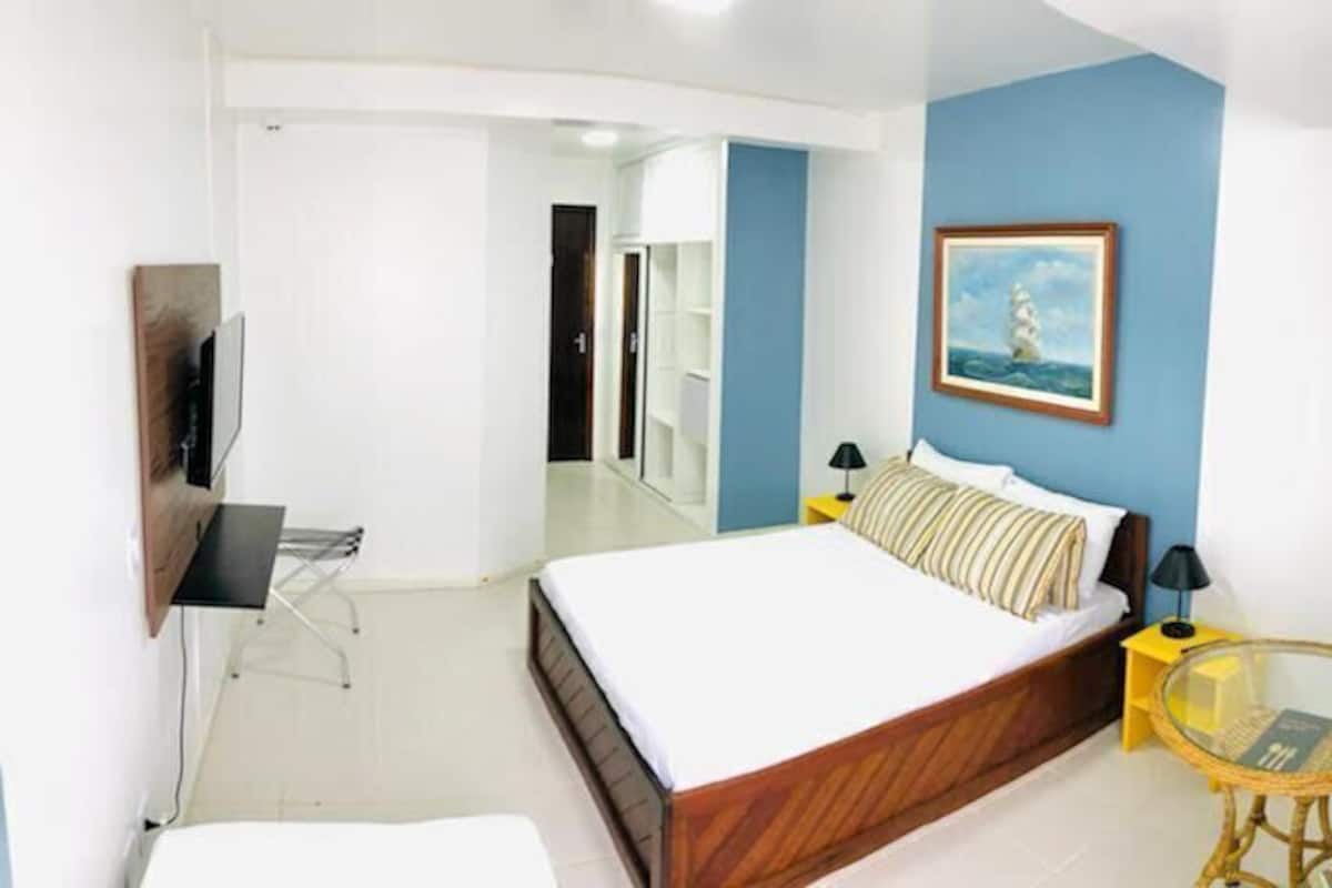A bedroom with a bed, a television, a chair, a small table, and a painting on the wall.