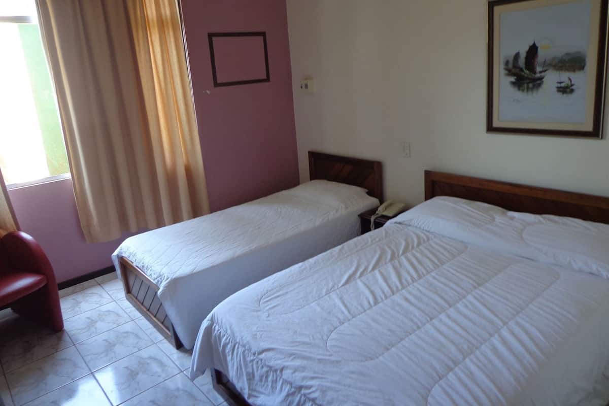 A room with two single beds, a chair, a bedside table with a phone, a framed picture on the wall, and a window with curtains.