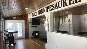A cozy living area with a fireplace, a dining table with chairs, and a sign reading "LAKE WINNIPEGUE". - Winnipesaukee - Lakefront house includes slip, deck, kayaks & sleeps up to 11 (Alton)