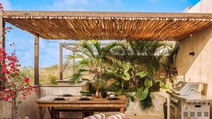 Un patio exterior con mesa de madera, banco y zona de barbacoa.