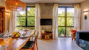 Un comedor con mesa de madera dispuesta para dos, un televisor sobre la chimenea y ventanas grandes con cortinas.
