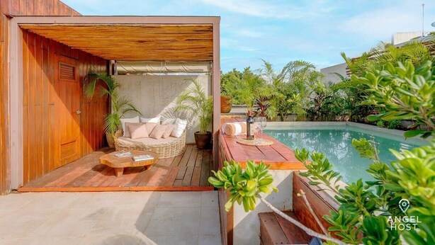 Una terraza de madera con un sofá de mimbre y una pequeña mesa, una piscina y abundante vegetación.
