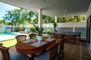 A covered outdoor dining area with a pool, wooden furniture, and tropical plants.