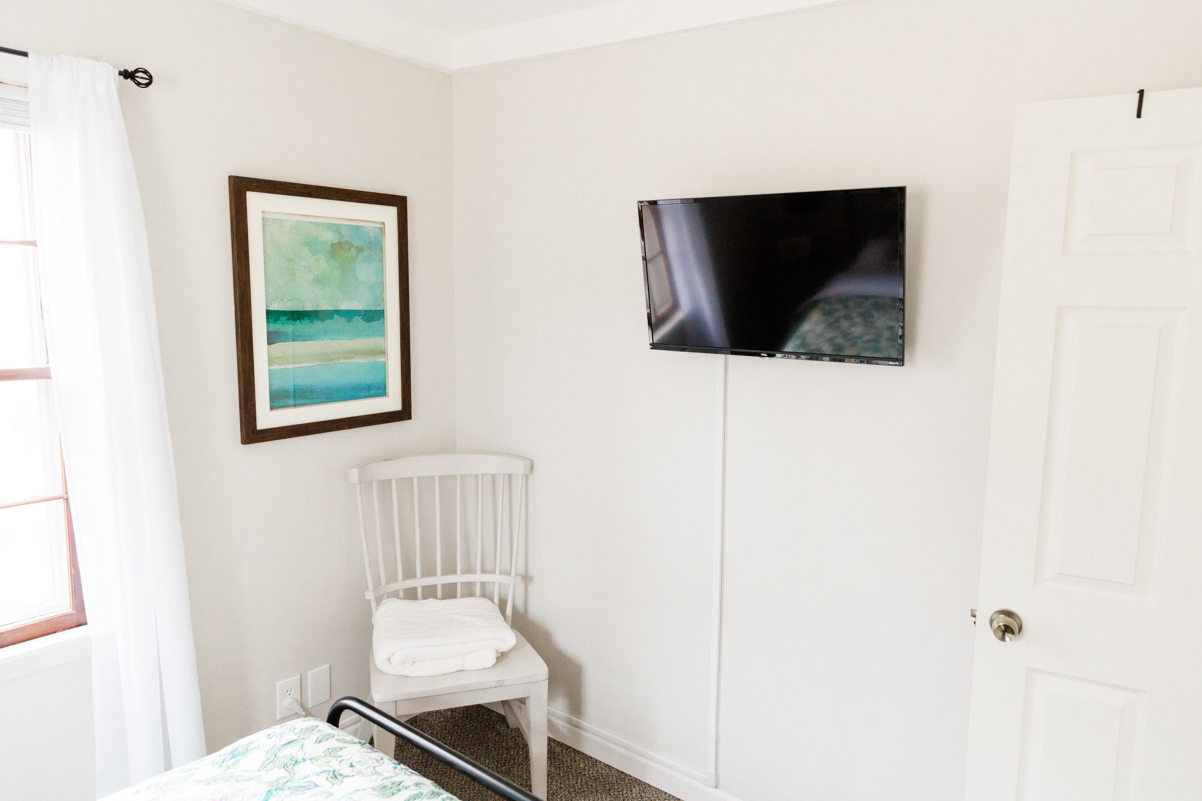 Un dormitorio con una cama, una silla blanca, un cuadro enmarcado y un televisor montado en la pared.