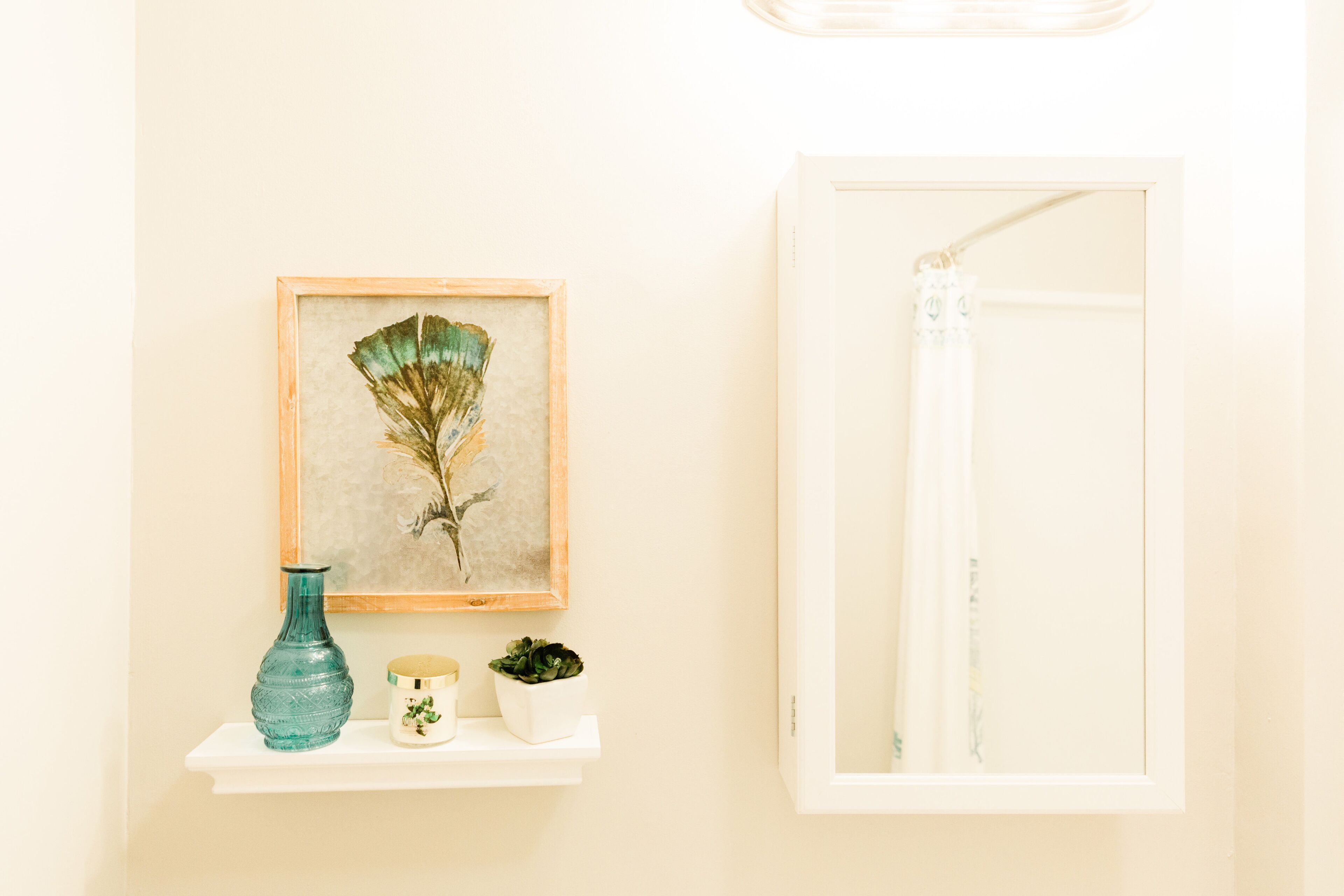 Une salle de bain avec une œuvre d’art encadrée, un vase turquoise, un petit récipient blanc et une étagère blanche.