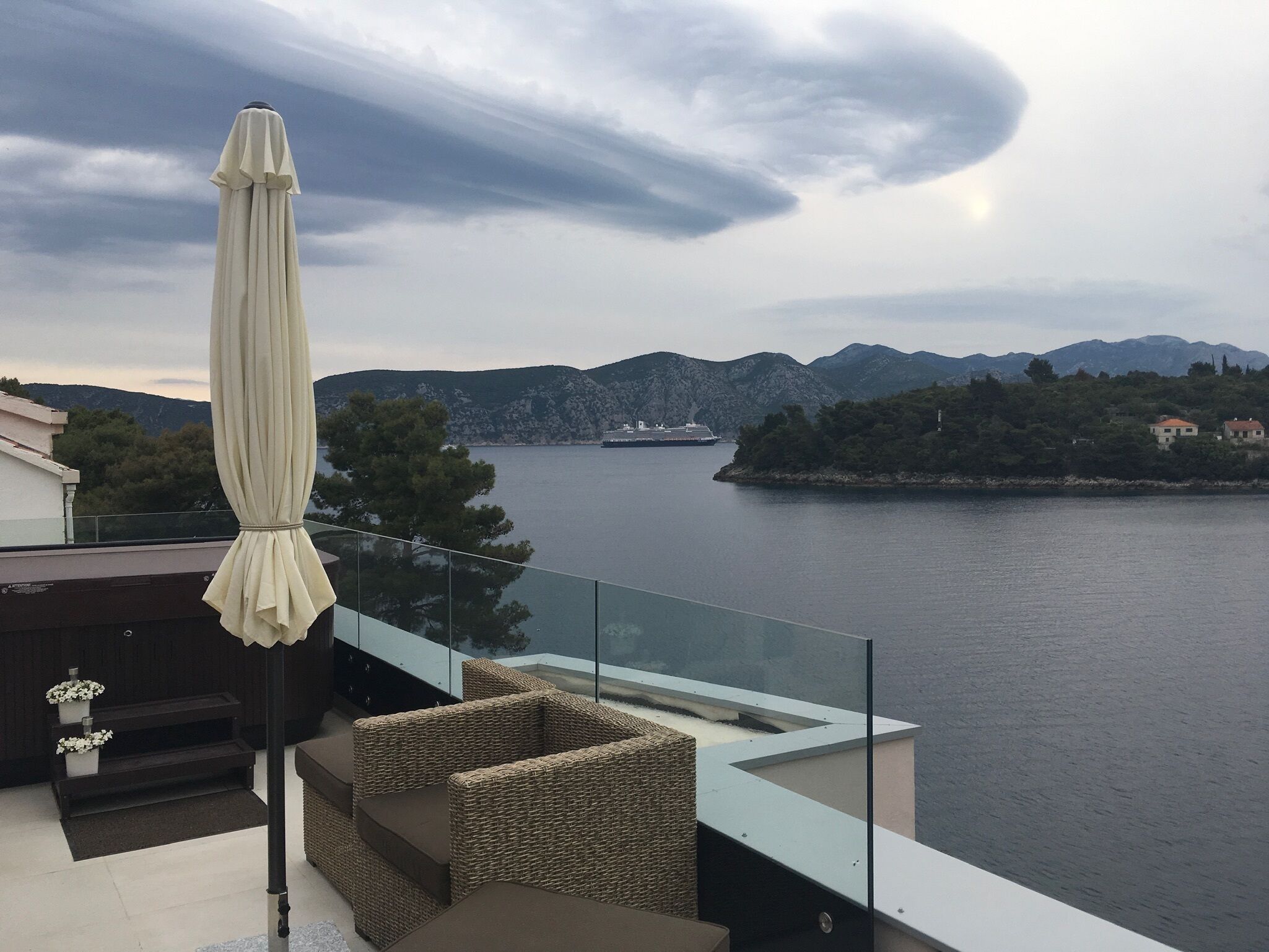 A balcony with a glass railing overlooking a body of water, a ship in the distance, and a mountainous landscape.