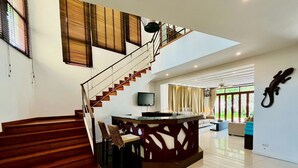 Uma sala de estar ampla com escada de madeira, ilha central com assentos, TV de tela plana e grandes janelas com vista para o verde.