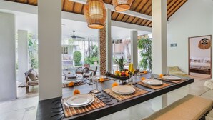 A dining area with a long table set for four, featuring woven placemats, plates, and cutlery. There are three woven pendant lights hanging above the table. The room has a high ceiling with exposed wooden beams and large windows that provide a view of the outdoor area. - Beautiful 4 Bedroom Villa, Oberoi (Seminyak)