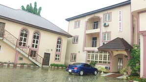 A building with a staircase leading to a door marked "HALL," a blue car parked in front, and a covered entrance with a tiled roof. - Chisam Suites Annex (Port Harcourt)