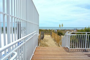 A wooden deck leading to a sandy beach area with a white fence and a view of the ocean.