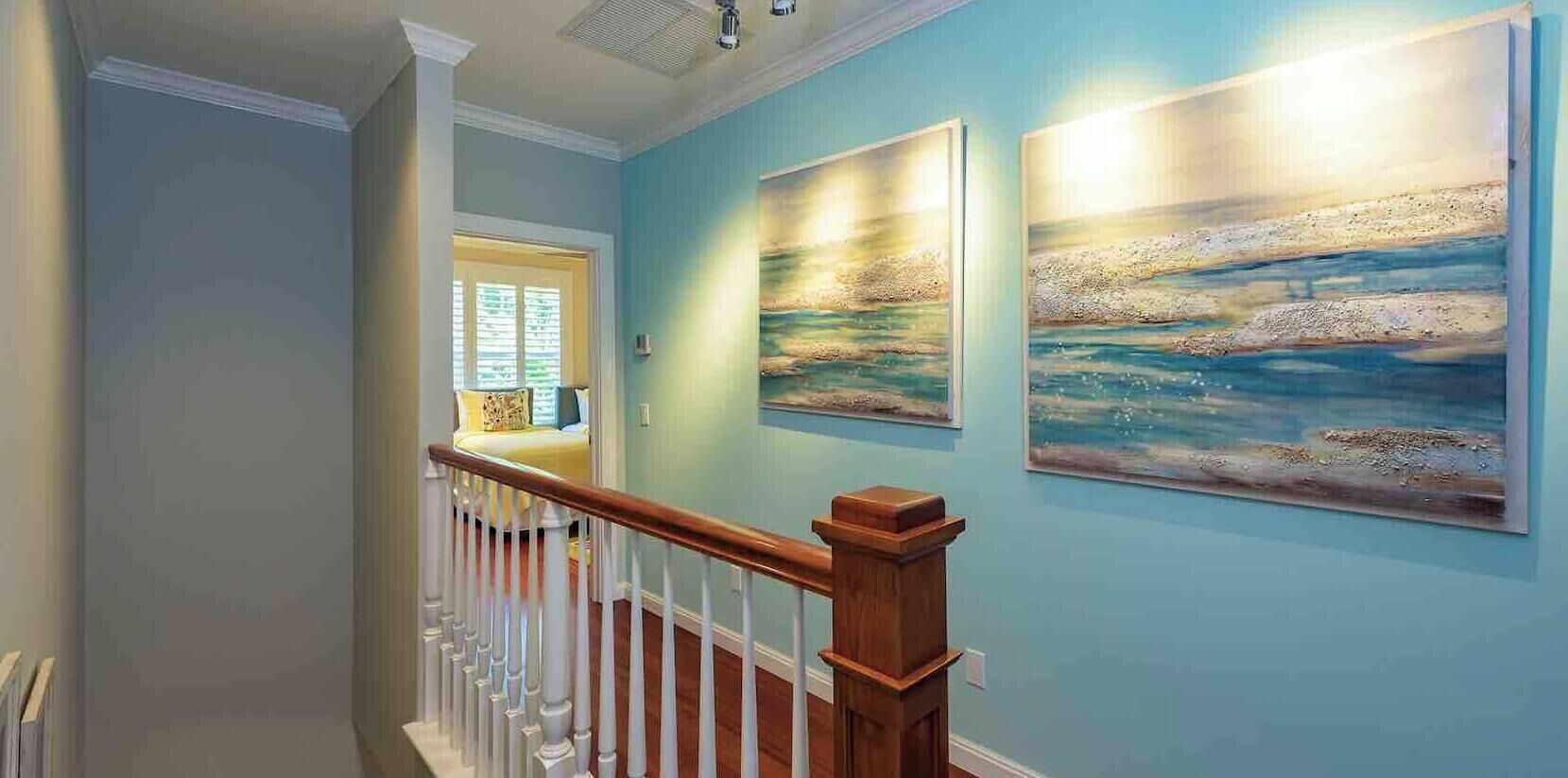 A staircase with white balusters and a wooden handrail leading to a room with light blue walls and two landscape paintings.