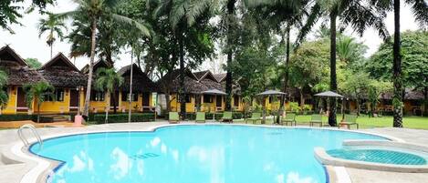 A swimming pool surrounded by palm trees and yellow buildings with thatched roofs.