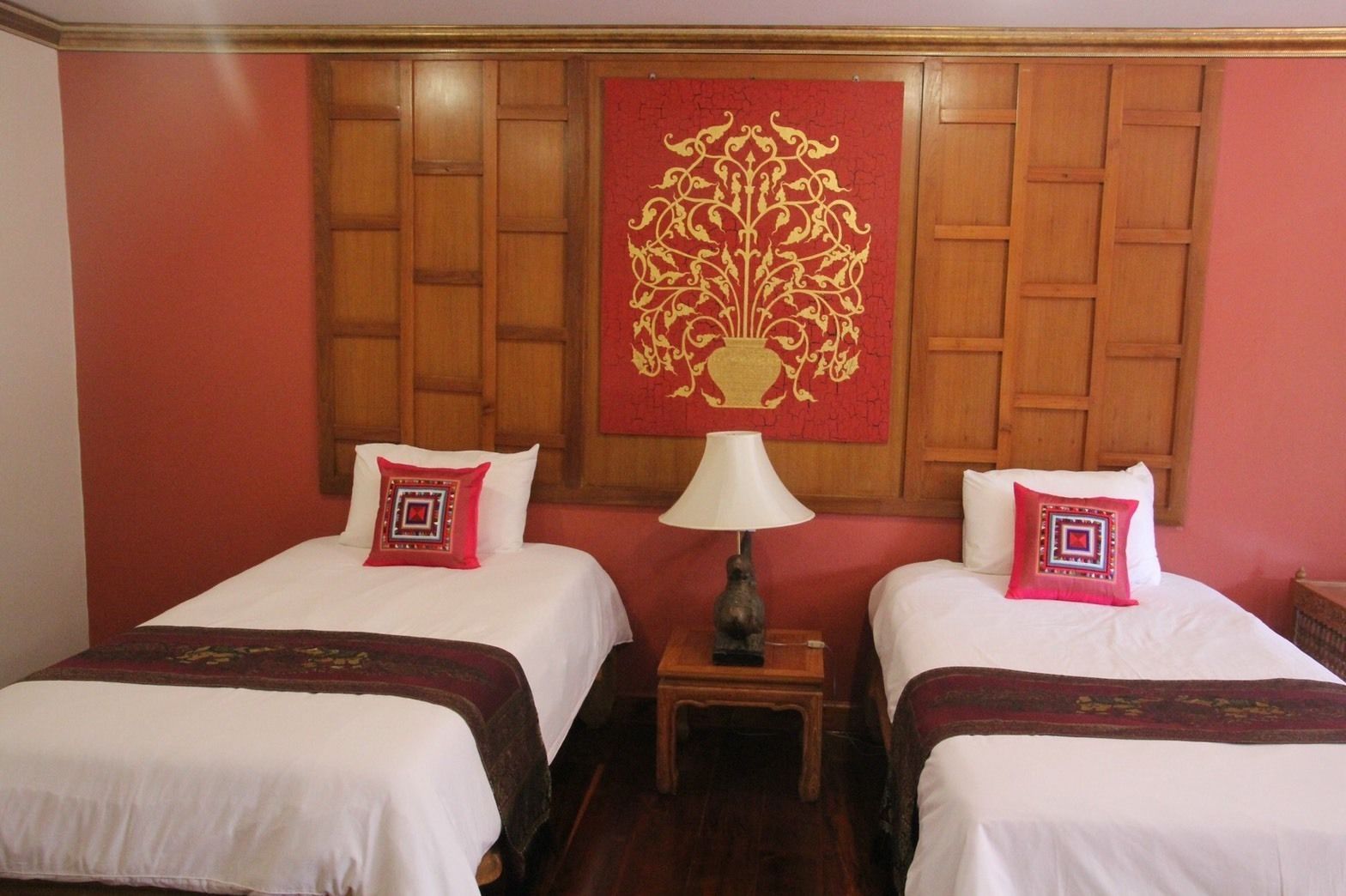 Habitación de hotel con dos camas, cada una con almohada roja y blanca, cabecero de madera y un tapiz decorativo rojo.