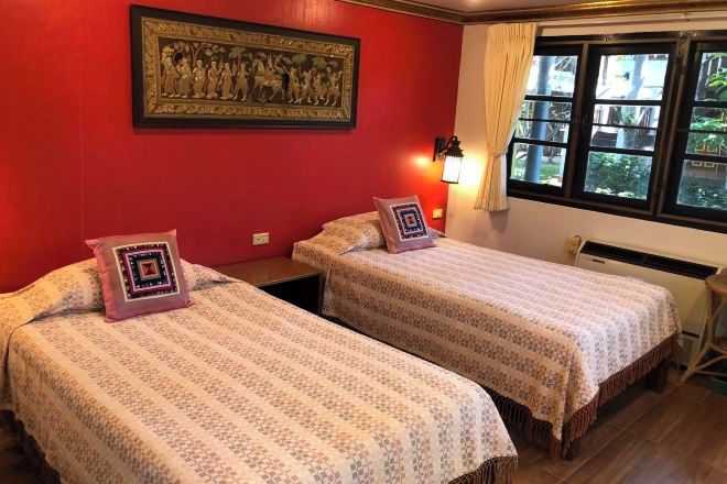 Une chambre d’hôtel avec deux lits, un mur rouge, une œuvre d’art encadrée, une fenêtre avec des rideaux et une table de chevet avec une lampe.
