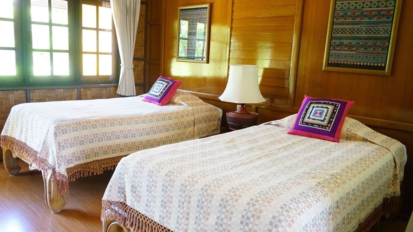 Two single beds with patterned bedding and decorative pillows, a bedside table with a lamp, and a window with curtains.